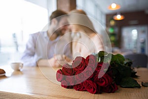 Bouquet of red roses on table at coffee shop and romantic young couple on background, selective focus