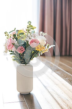 A bouquet of roses, ranunculus, and yellow blossoms graces a white ribbed vase, with pink curtains providing a soft and enchanting