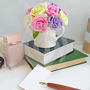 Bouquet of roses in a vase, stack of books, card with mountain pen on table in front of white background.