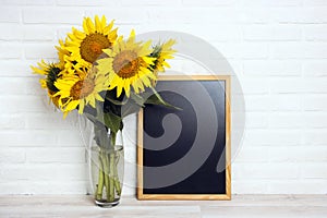 A bouquet of sunflowers in a vase and empty chalkboard against the background of a white brick wall