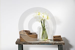 Bouquet of three white lilies in a tall glass vase on a beige vintage bench against a gray wall. Wooden boxes of different sizes