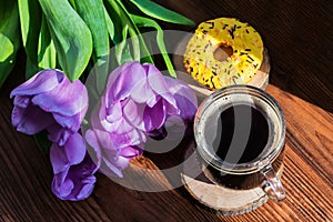 A bouquet of tulips with lilac petals lies on a wooden table next to a mug of black coffee and a donut