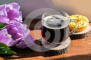 A bouquet of tulips with lilac petals lies on a wooden table next to a mug of black coffee and a donut
