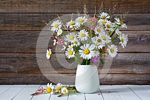 Bouquet of white daisies and other wildflowers in a white vase on a wooden background