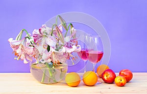 Bouquet of white lilies with purple spots in glass round vase, glasses of rose wine and fresh fruits on wooden table. Purple