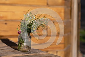 Bouquet of white and pink wildflowers in glass jar is placed on wooden table near wall of house
