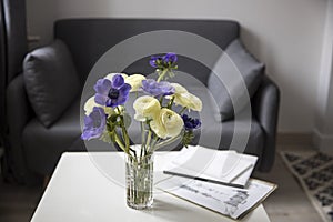 Bouquet of white ranunculus and blue anemone in the vase on a white coffee table near sofa with plaid. The books and journal