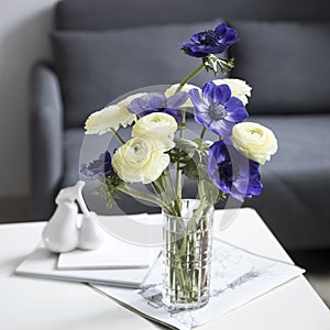 Bouquet of white ranunculus and blue anemone in the vase on a white coffee table near sofa with plaid. The books and journal