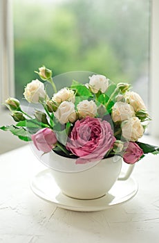 A bouquet of white roses in a cup on a white table opposite the window