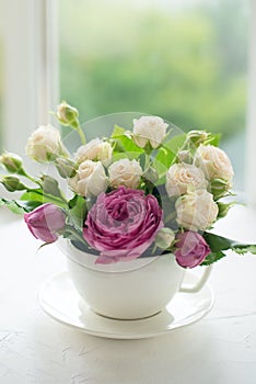 A bouquet of white roses in a cup on a white table opposite the window