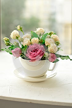 A bouquet of white roses in a cup on a white table opposite the window