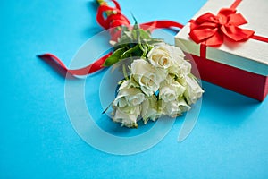 Bouquet of white roses with red bow on blue background. Boxed gift on side