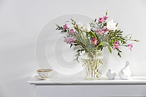 A bouquet of white tulips, pink eustoma, hyacinth, eucalyptus in fluted glass vase on panel of an artificial fireplace. Tea in a
