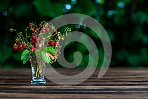 bouquet wild strawberry in transparent shot glass stays on the brown wooden table with green leafs on the back, with placeholder