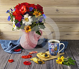 A bouquet of wildflowers with red poppies, a blue cup of coffee and poppy seed cookies on a wooden background