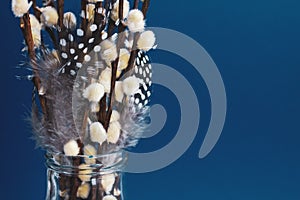 A bouquet of willow and bird feathers stands in a transparent glass vase on a blue background. copy space for text.