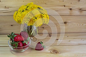 A bouquet of yellow dandelions on a warm wooden background. red strawberries in a glass vase.