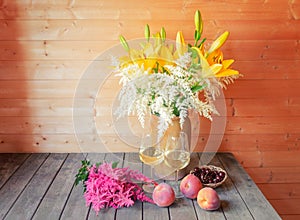 Bouquet of yellow lilies and white astilbe in clay vase, glasses of white wine, pink astilbe and fresh fruits near