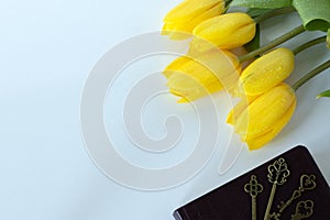 Bouquet of yellow tulips with ancient keys placed on closed holy bible book on white background, top view