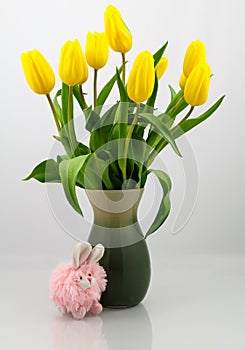 Bouquet of yellow tulips in a pleasing green vase isolated on a pale background. Pink bunny accents the base of the vase.