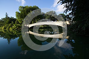 Bow bridge, trees and blue sky reflect on calming water at Central Park