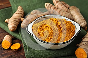 Bowl with aromatic turmeric powder and cut roots on wooden table, closeup