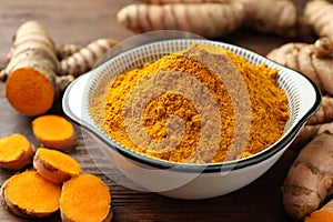 Bowl with aromatic turmeric powder and cut roots on wooden table, closeup