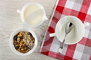 Bowl with baked muesli, pitcher with yogurt, spoon in bowl on napkin on wooden table. Top view