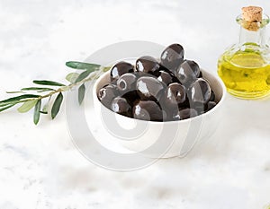 A bowl of black olives sits on a table next to a bottle of olive oil