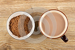 Bowl with cacao powder, brown cup of cocoa with milk on table. Top view