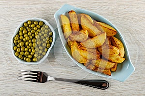 Bowl with green peas, blue plate with fried potato, fork on wooden table. Top view