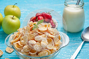 Bowl of cereal on a blue wooden table and fresh strawberry, apple, milk behind.