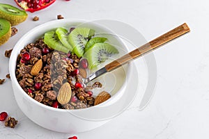 Bowl of chocolate oat granola with yogurt, fresh kiwi fruit, pomegranate seeds, nuts and spoon on white marble table