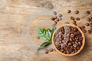 Bowl of coffee beans and fresh green leaves on wooden table, top view
