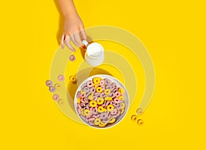 Bowl of colorful cereal corn rings on yellow table. Breakfast concept. Kid hand pouring milk into bowl