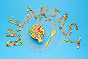 A bowl with colorful corn sticks, a yellow spoon and the inscription breakfast on a blue background. Flat lay