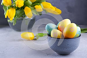 Bowl with colorful Easter eggs; spring easter decoration on gray table with bouquet of yellow tulip flowers in glass vase