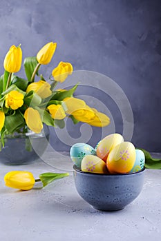 Bowl with colorful Easter eggs; spring easter decoration on gray table with bouquet of yellow tulip flowers in glass vase;