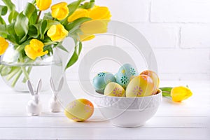 Bowl with colorful Easter eggs, spring easter decoration on white wooden table with bouquet of yellow tulip flowers in glass vase
