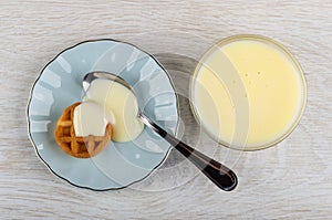 Bowl with condensed milk, round waffle, teaspoon with condensed milk in saucer on wooden table. Top view