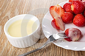 Bowl with condensed milk, strawberries in plate, berry strung on fork on wooden table