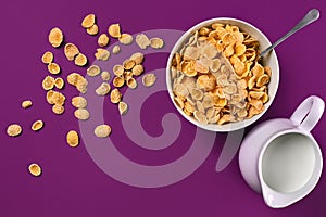 Bowl with corn flakes, jug of milk and spoon on purple background, top view