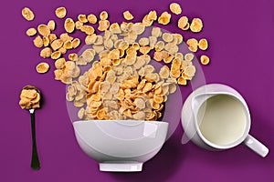 Bowl with corn flakes, jug of milk and spoon on purple background, top view