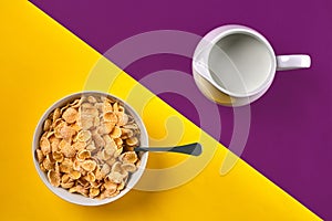 Bowl with corn flakes, jug of milk and spoon on purple and yellow background, top view
