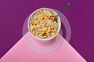 Bowl with corn flakes and spoon on purple and pink background, top view