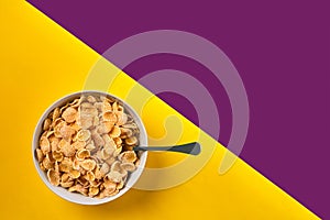 Bowl with corn flakes and spoon on purple and yellow background, top view