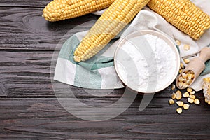 Bowl with corn starch, ripe cobs and kernels on dark wooden table, flat lay