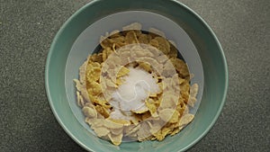 In a bowl of cornflakes, 2 spoons of sugar are added, seen in close-up top view