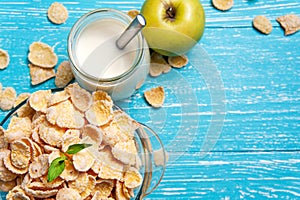 Bowl of cornflakes cereal on a blue wooden table and fresh apple, milk behind.