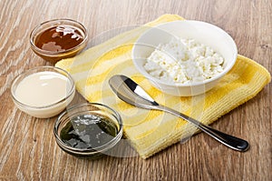 Bowl with cottage cheese, spoon on napkin, bowls with boiled condensed milk, condensed milk, jam on table